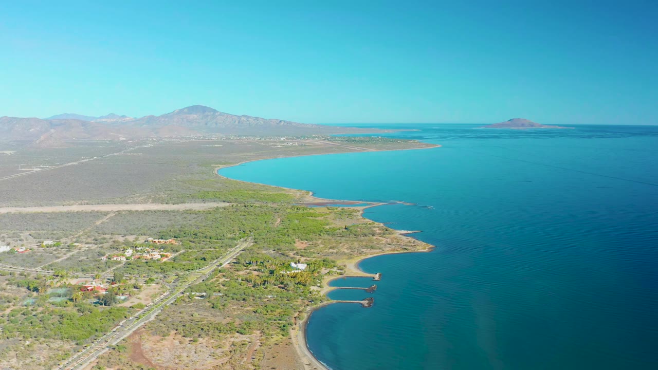 High coastal view with blue water, curving shoreline, road, vegetation, buildings, mountains, and clear sky.