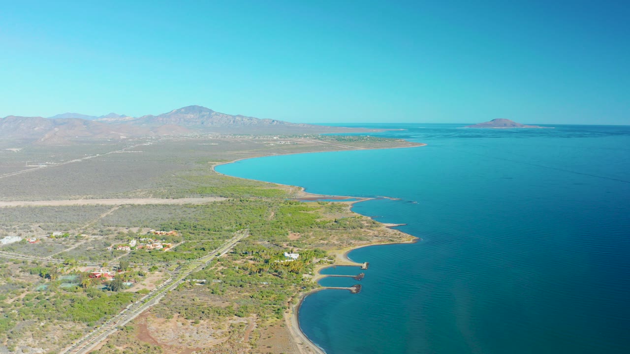Aerial view over Loreto, Baja California Sur, with turquoise water on the right, desert vegetation and a straight road or runway at left, distant mountains under clear sky, and no visible people.