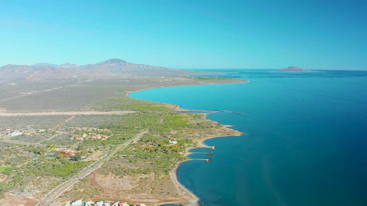 Aerial frame over Loreto, Baja California Sur showing a coastal road, scattered buildings, curved shoreline, bright blue water, distant mountains, and no visible people.