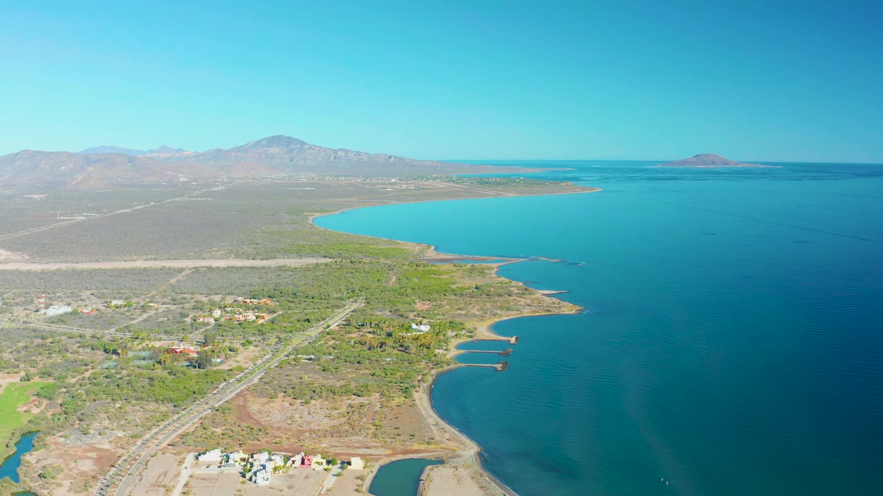 Aerial frame over Loreto showing the road running near a curved shoreline, small buildings in dry terrain, blue coastal water, mountains on the horizon, and no visible people.