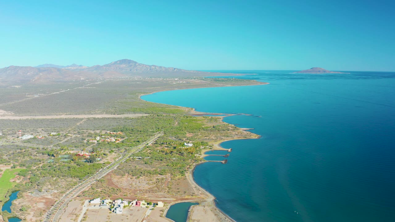 Loreto aerial view with the road crossing the lower left, scattered structures near dry lots, a hooked shoreline, bright blue water to the right, distant mountains, and no visible people.