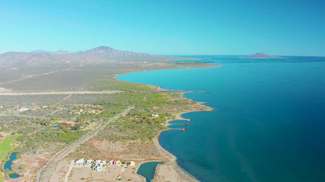 Wide aerial of road traffic beside a curving shoreline and arid land.