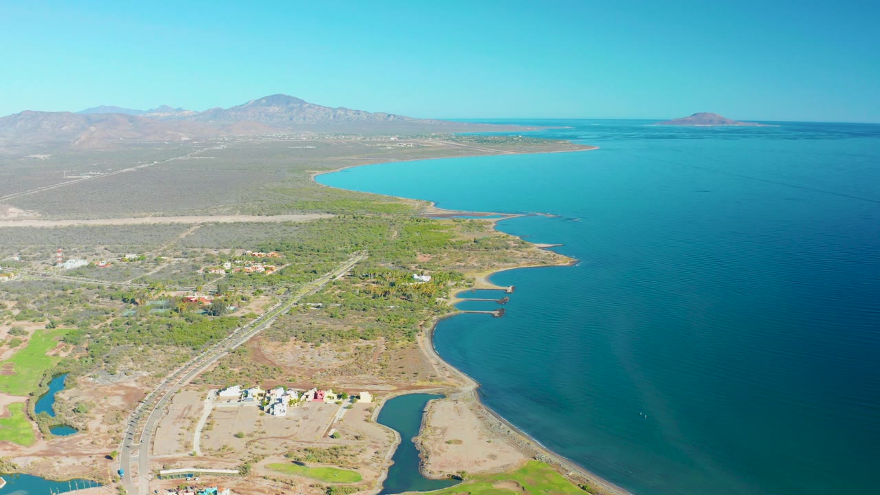 Opening aerial view over Loreto, Baja California Sur, showing blue coastal water, a curving shoreline, dry land, scattered buildings, a road, distant mountains, and no visible people.