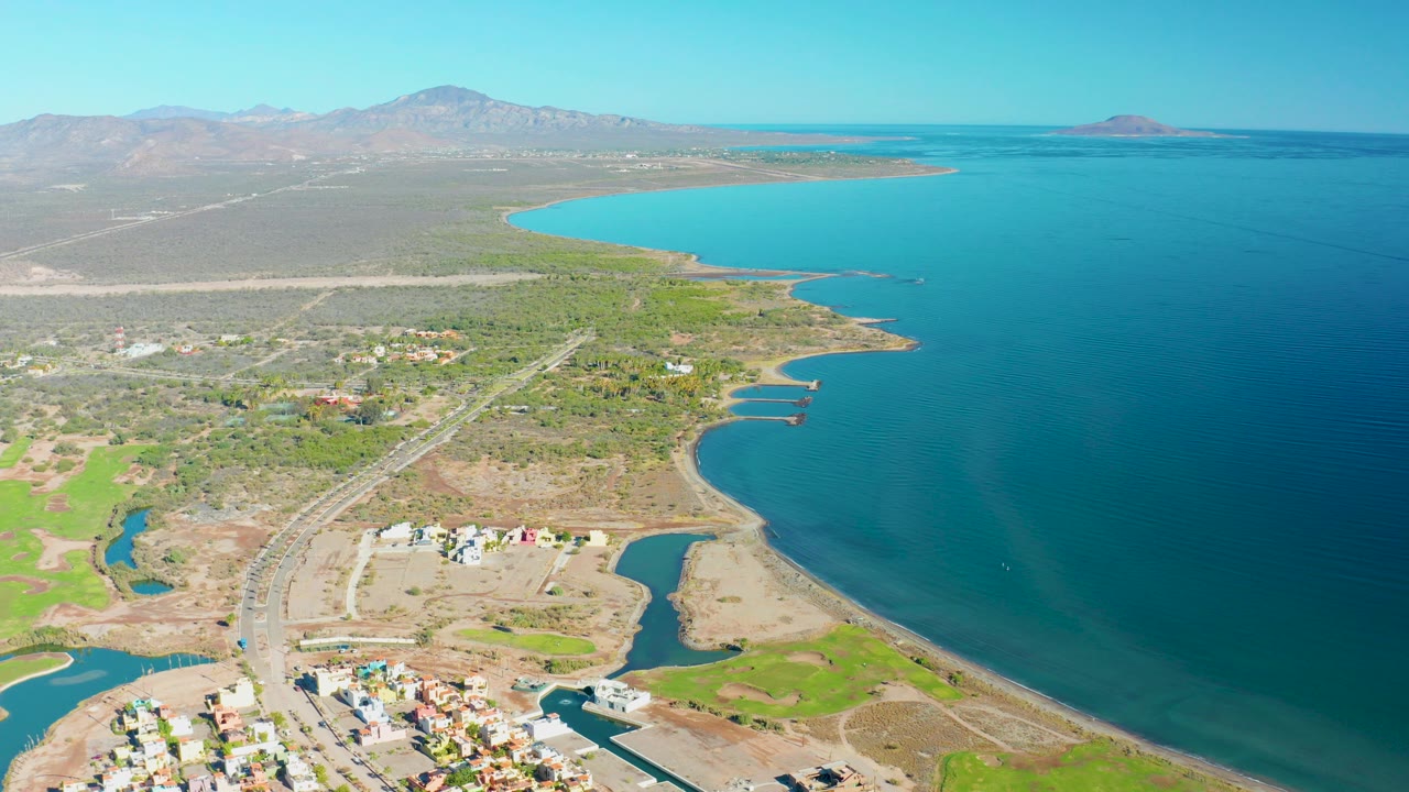 Opening aerial frame over Loreto, Baja California Sur shows turquoise bay water beside exposed tan shoreline, a small settlement, green fields, a road, distant mountains, and no visible people.