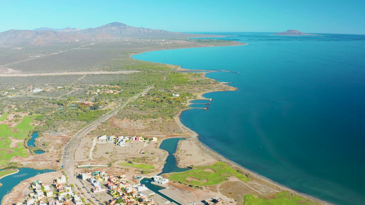 At 0:01, the Loreto aerial view holds on the curved bay, with the settlement low in frame, a road crossing the dry and green terrain, mountains beyond, and no visible people.