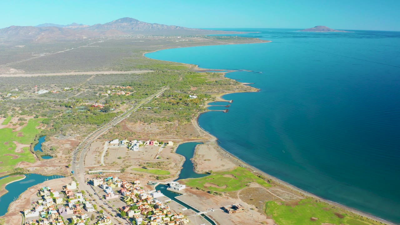 Drone view over shoreline with road, settlement edge, green fields, dry terrain, bay, and mountains.