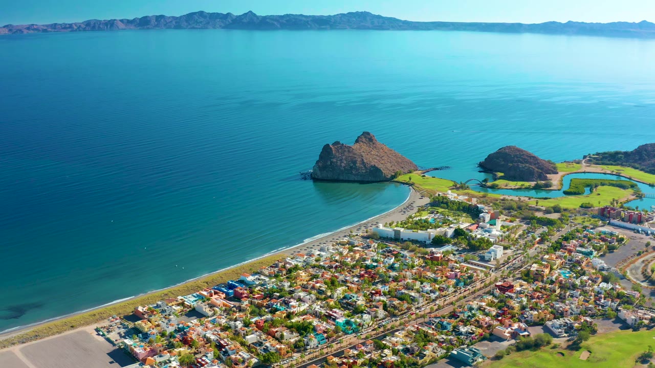 Opening aerial frame over Loreto, Baja California Sur, with turquoise ocean above a coastal town, a curved beach, rocky offshore formations, distant mountains, and no visible people.