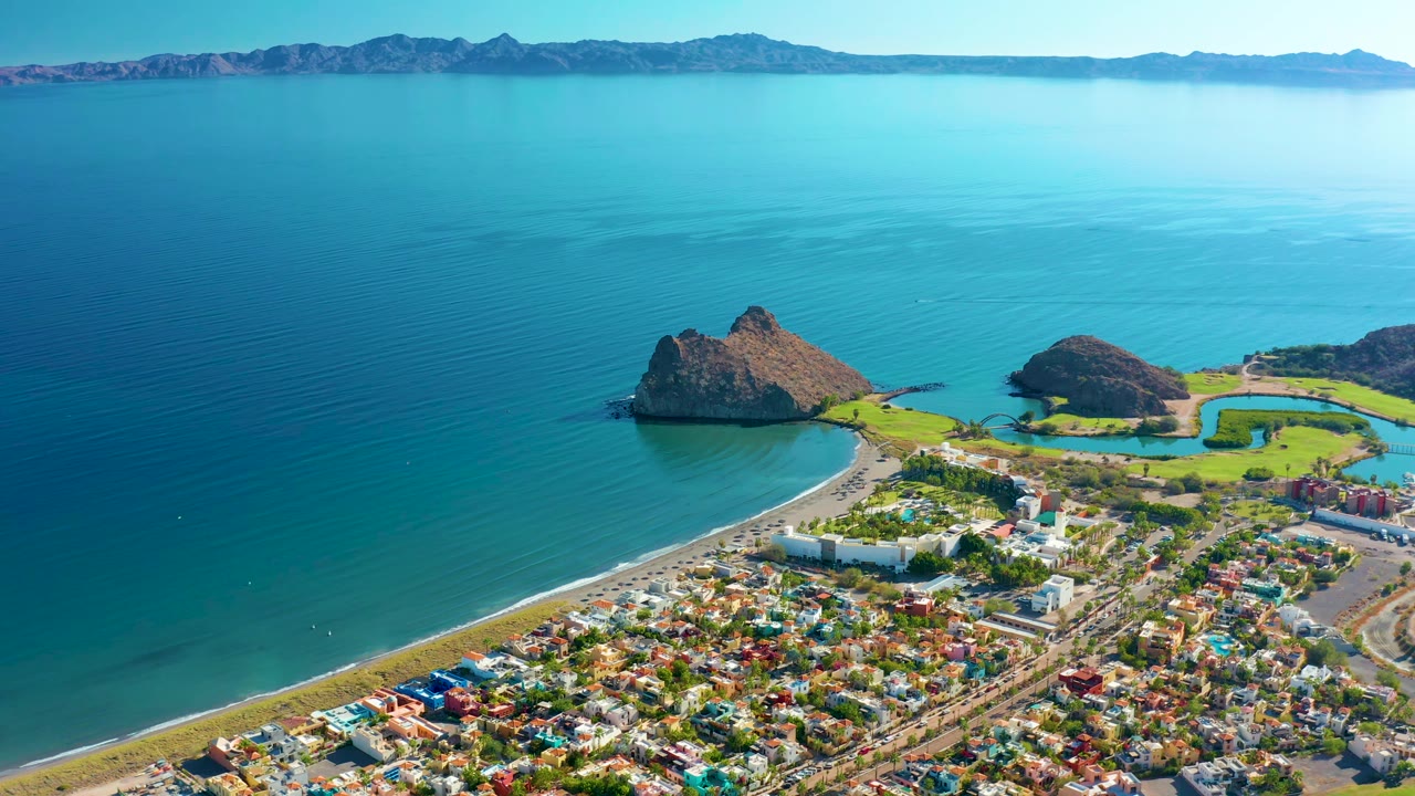 Aerial view over Loreto showing more of the curved shoreline and town grid below, with rippled blue water to the left, rocky outcrops near the coast, distant mountains, and no visible people.