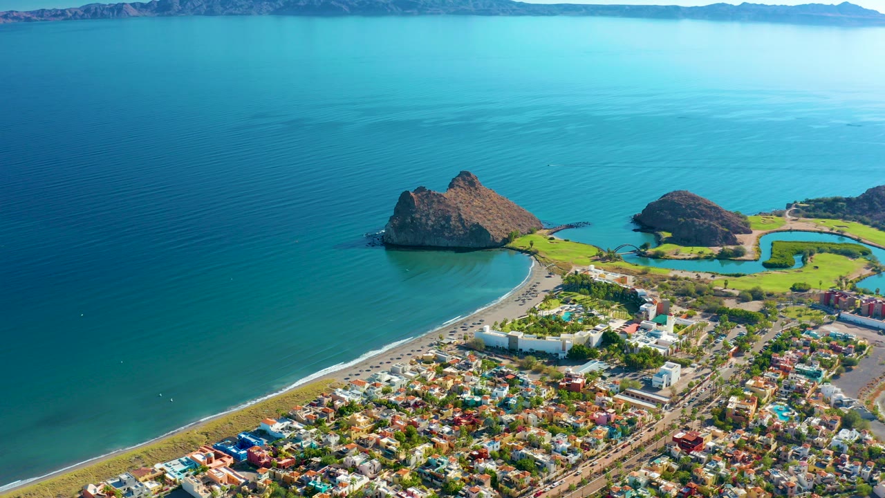 Opening aerial frame over Loreto, Baja California Sur, showing the town at the bottom edge, a curved beach, turquoise ocean, rocky coastal landforms, distant mountains, and no visible people.