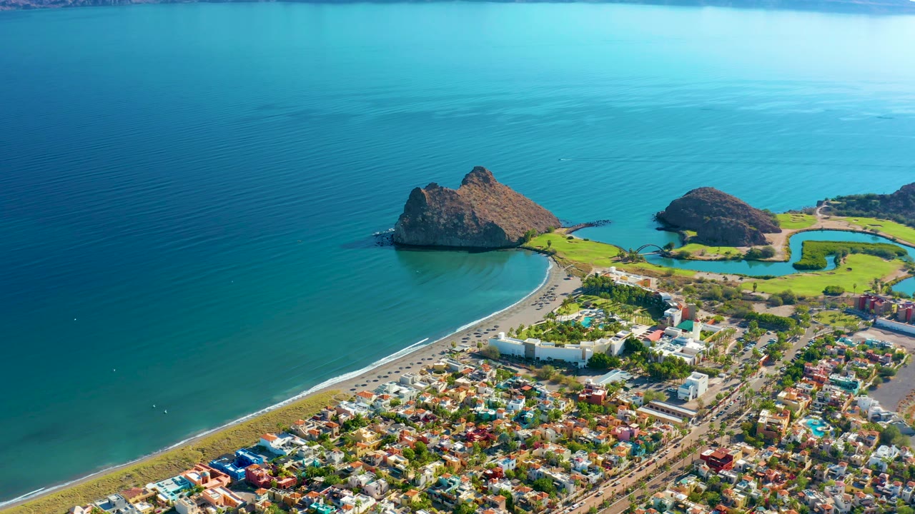 Wide aerial coastline with beach, town, bright blue water, and mountains on the horizon.