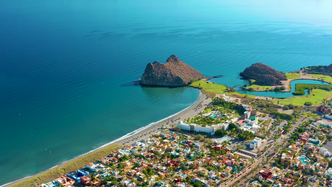 Aerial frame over Loreto, Baja California Sur, showing turquoise ocean, two rocky outcrops, a curved beach, shoreline road, colorful town blocks, and no visible people.