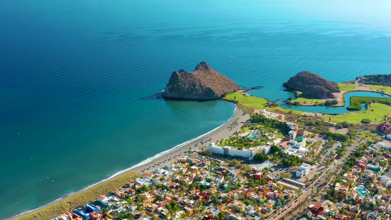 Drone-style view of beach, resort-like buildings, town blocks, and ocean rocks.