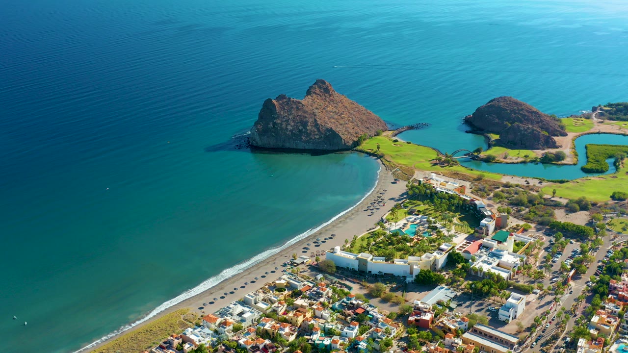 Aerial view over Loreto, Baja California Sur, with turquoise ocean, a curved sandy beach, roadside buildings, green grounds, rocky offshore landforms, and no visible people.