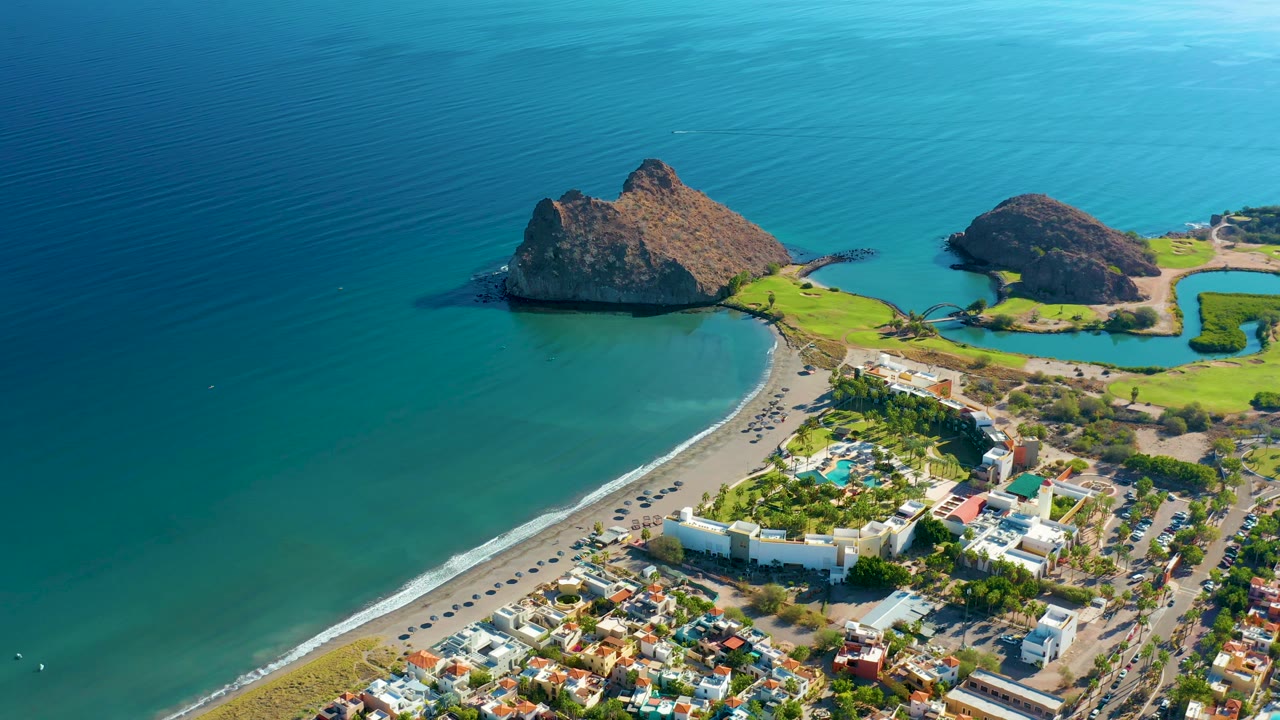 Drone-style coastal view of turquoise water, sandy shoreline, road traffic, greenery, and buildings.