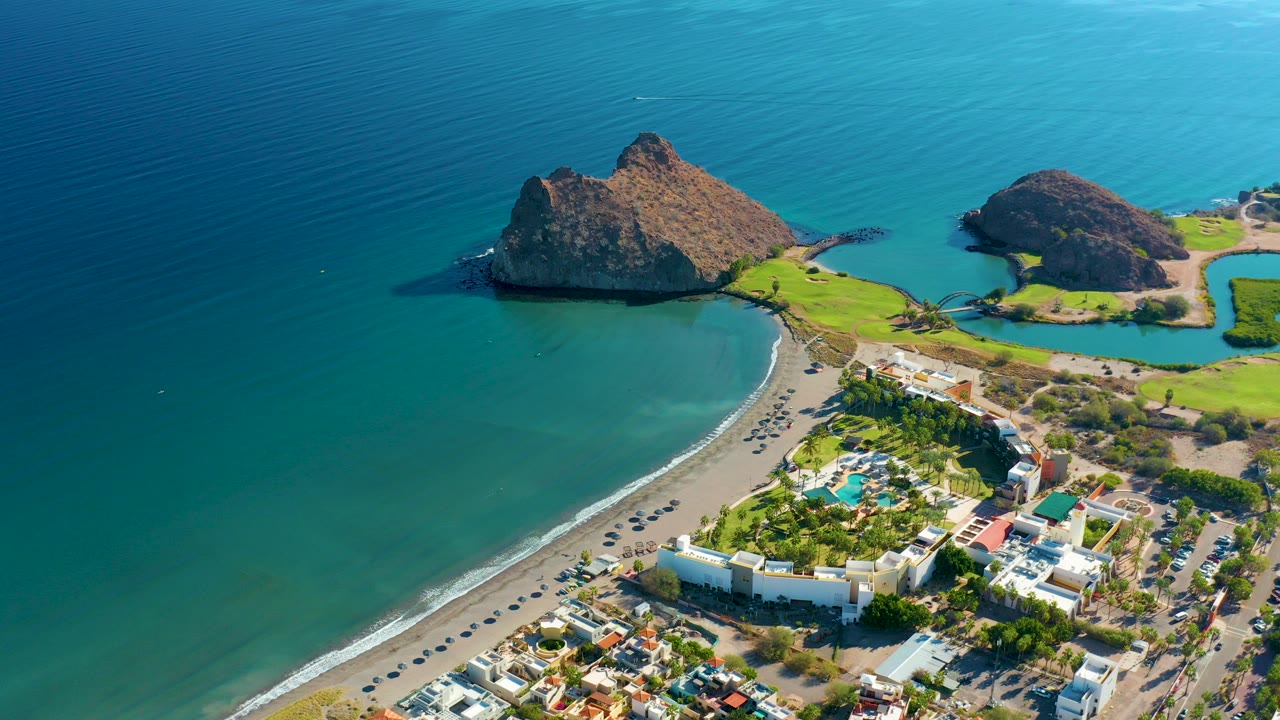 Aerial view of curved beach, rocky outcrops, turquoise water, and coastal buildings.