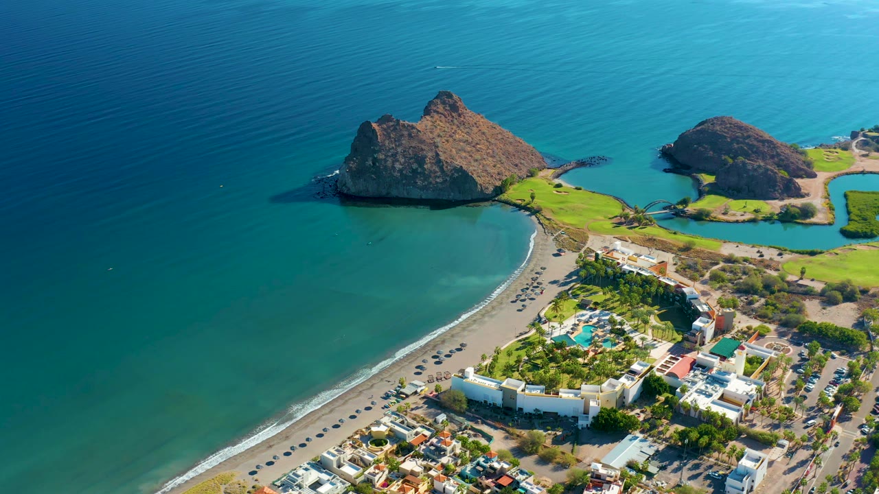 At 0:01, the Loreto aerial view holds on the same shoreline, with the larger rocky outcrop casting dark shadow into the water and the beach road and developed grounds visible below, with no visible people.