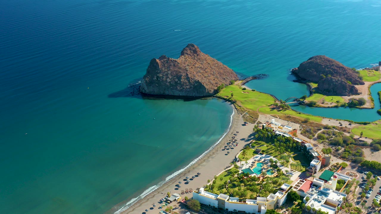 Aerial frame over Loreto, Baja California Sur showing a curved sandy beach, resort pools and buildings, green lawns, rocky headlands, turquoise shallows, and tiny people on the sand.