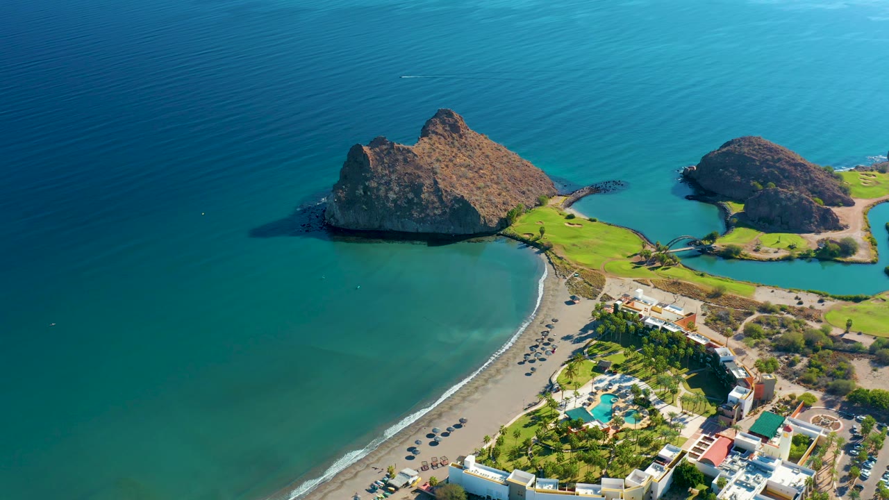 The 0:02 frame looks down on Loreto's sandy shoreline with a broader sweep of blue water, rocky headlands near the top edge, resort roofs and pools below, and small people scattered on the beach.