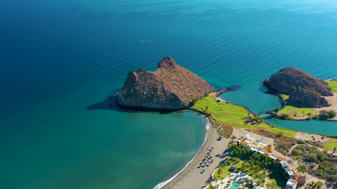 Opening aerial frame over Loreto, Baja California Sur, with two rocky islands, a curved beach, green shoreline grass, parked vehicles, small buildings, and no visible people.