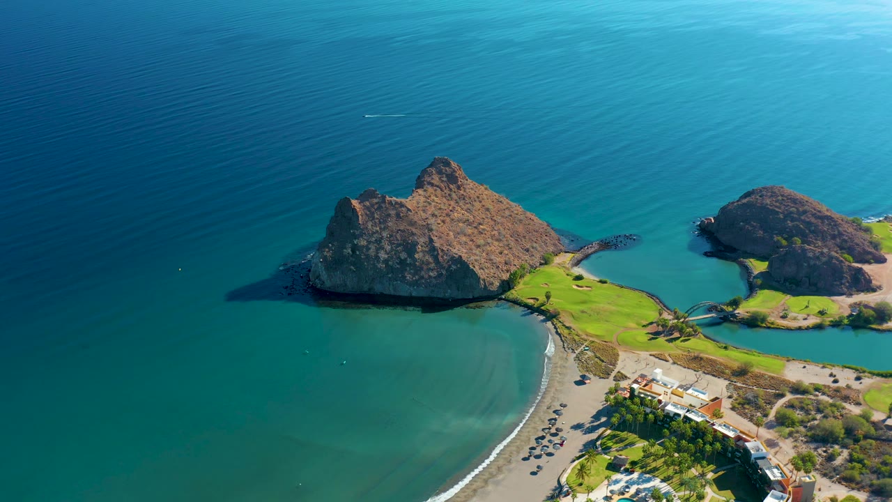 Aerial frame over Loreto, Baja California Sur, showing rocky islands in blue water, the crescent beach at lower left, green grass and a shoreline road at lower right, and no visible people.
