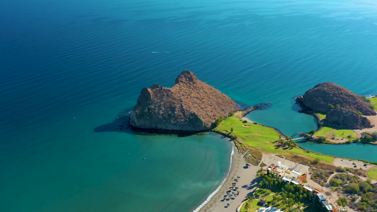 Aerial shot of a rocky island beside a sheltered beach and parking lot.
