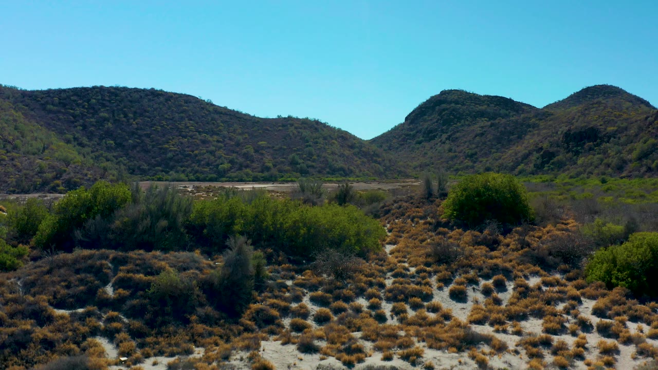 A wider Loreto, Baja California Sur valley view shows scrub-covered foreground, sandy washes, green vegetation, distant hills, and no visible people.