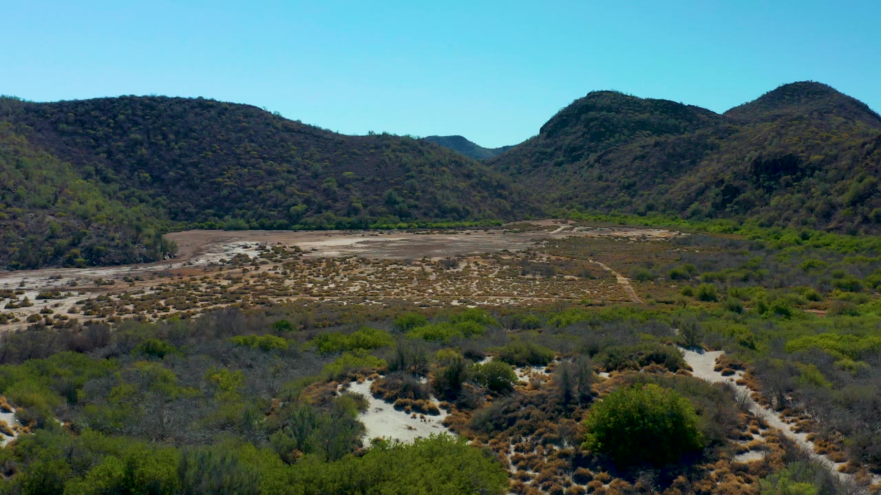 Wide aerial view of a dry Loreto, Baja California Sur valley with scrubby foreground, a pale riverbed, rounded mountain slopes, clear blue sky, and no visible people.