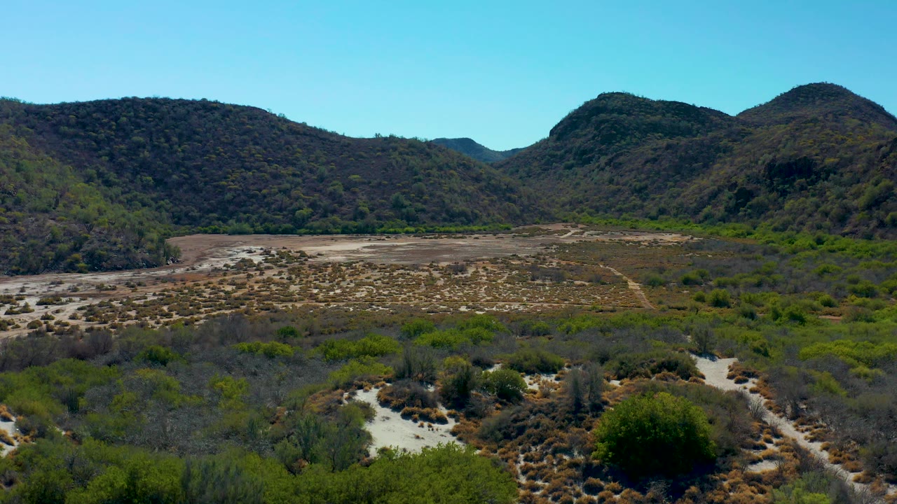 Aerial frame in Loreto, Baja California Sur showing a pale dry wash cutting through green shrubs toward dark brown mountain ridges, with no visible people.