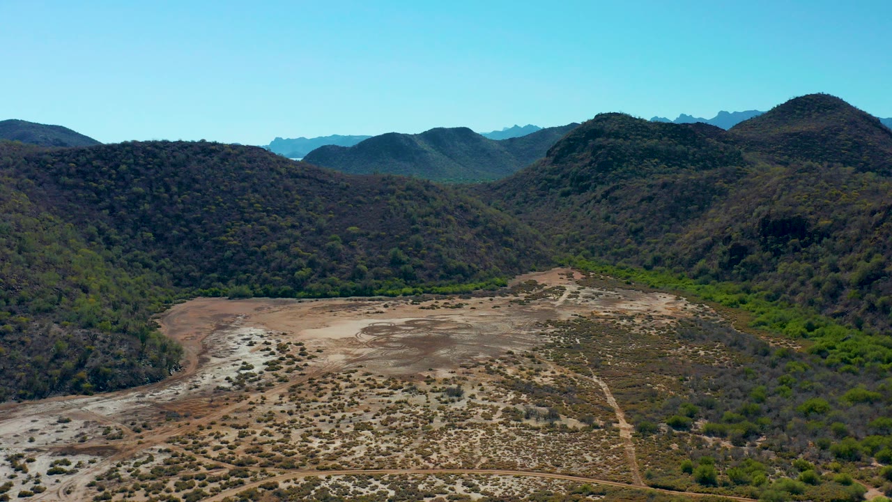 Opening aerial frame over a dry valley in Loreto, Baja California Sur, with scrubby tan ground, green hills, distant mountain layers, clear blue sky, and no visible people.