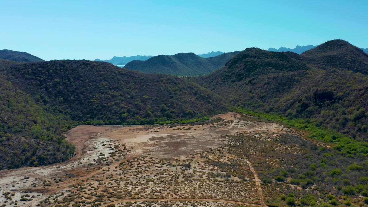 Aerial frame from Loreto, Baja California Sur, with rocky tan valley terrain in front, rounded green hills in the center, a small band of blue water beyond the mountains, and no visible people.