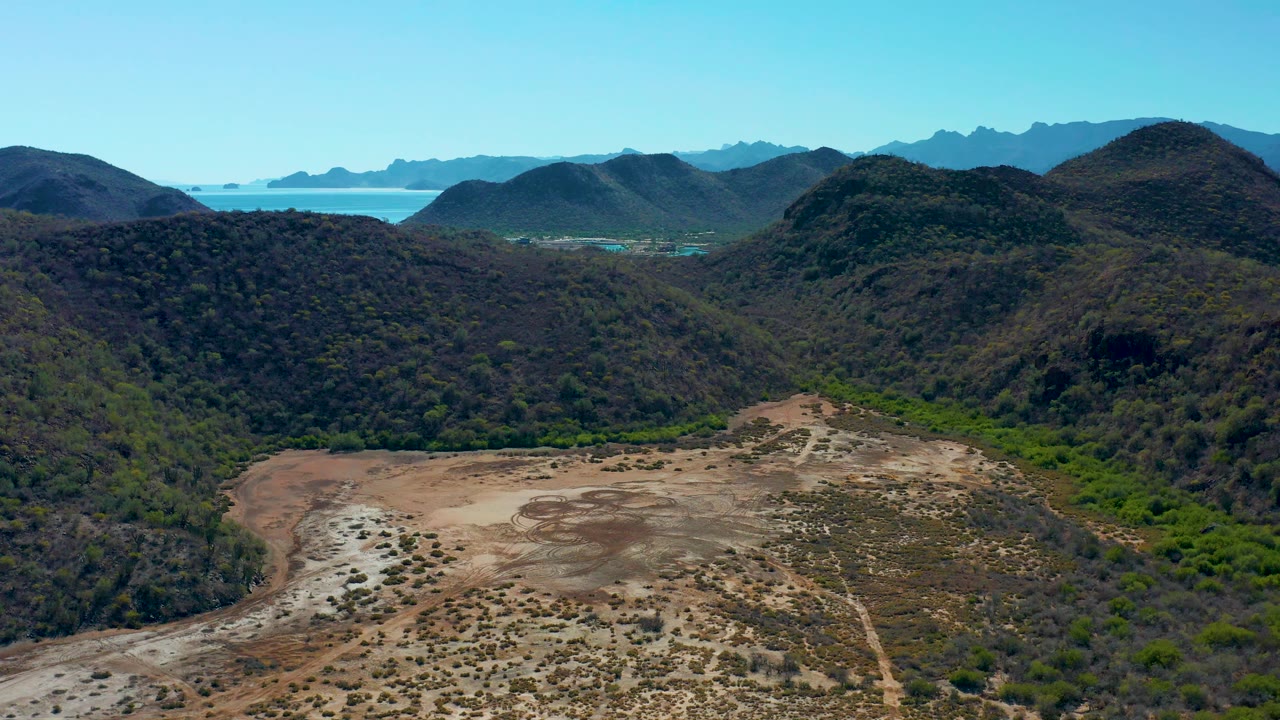 Aerial frame from Loreto, Baja California Sur showing pale desert ground with scattered shrubs, darker green drainage vegetation, rounded hills, and distant blue water, no visible people.