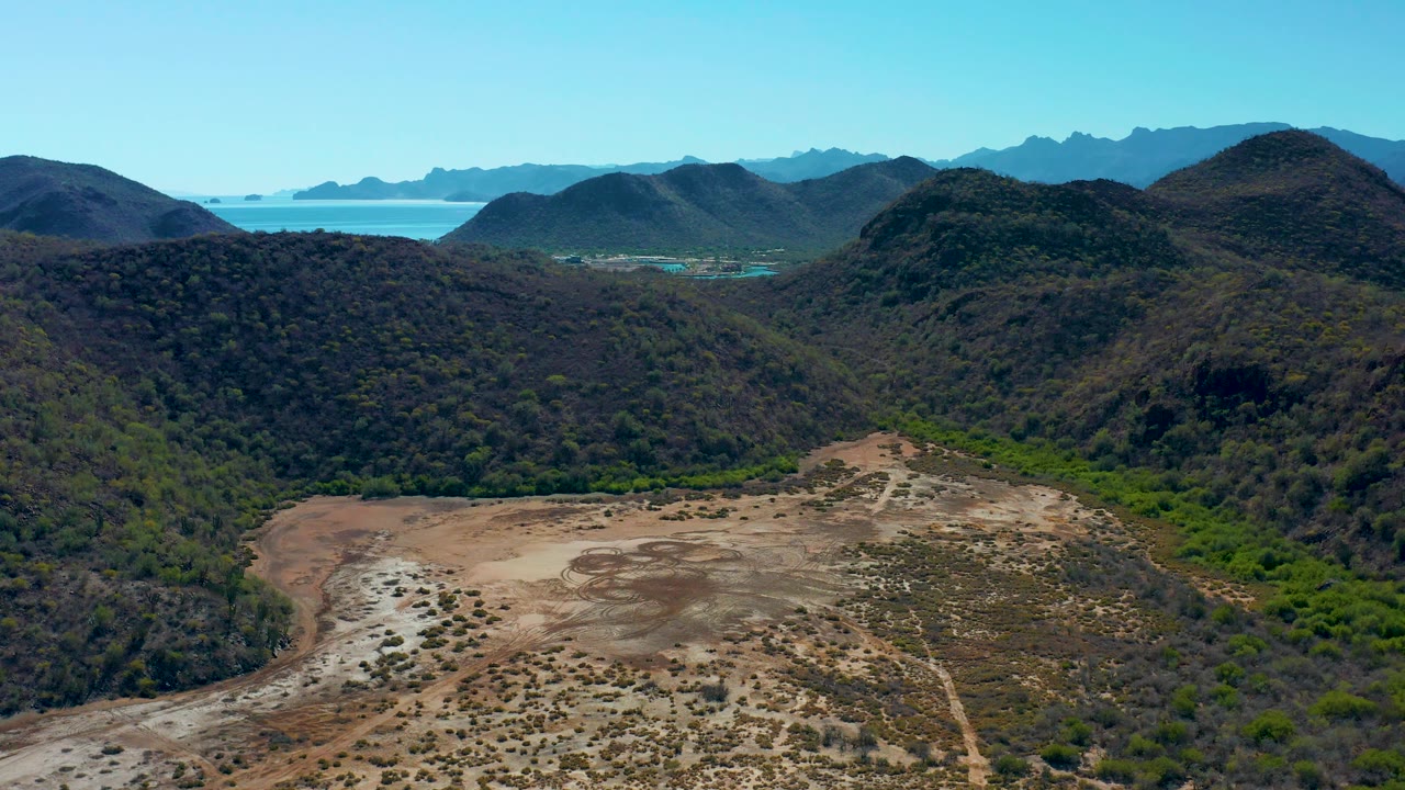 Aerial view over Loreto, Baja California Sur with a sandy basin in the foreground, scrub-covered hills in the middle distance, and a strip of ocean below mountain silhouettes, no visible people.