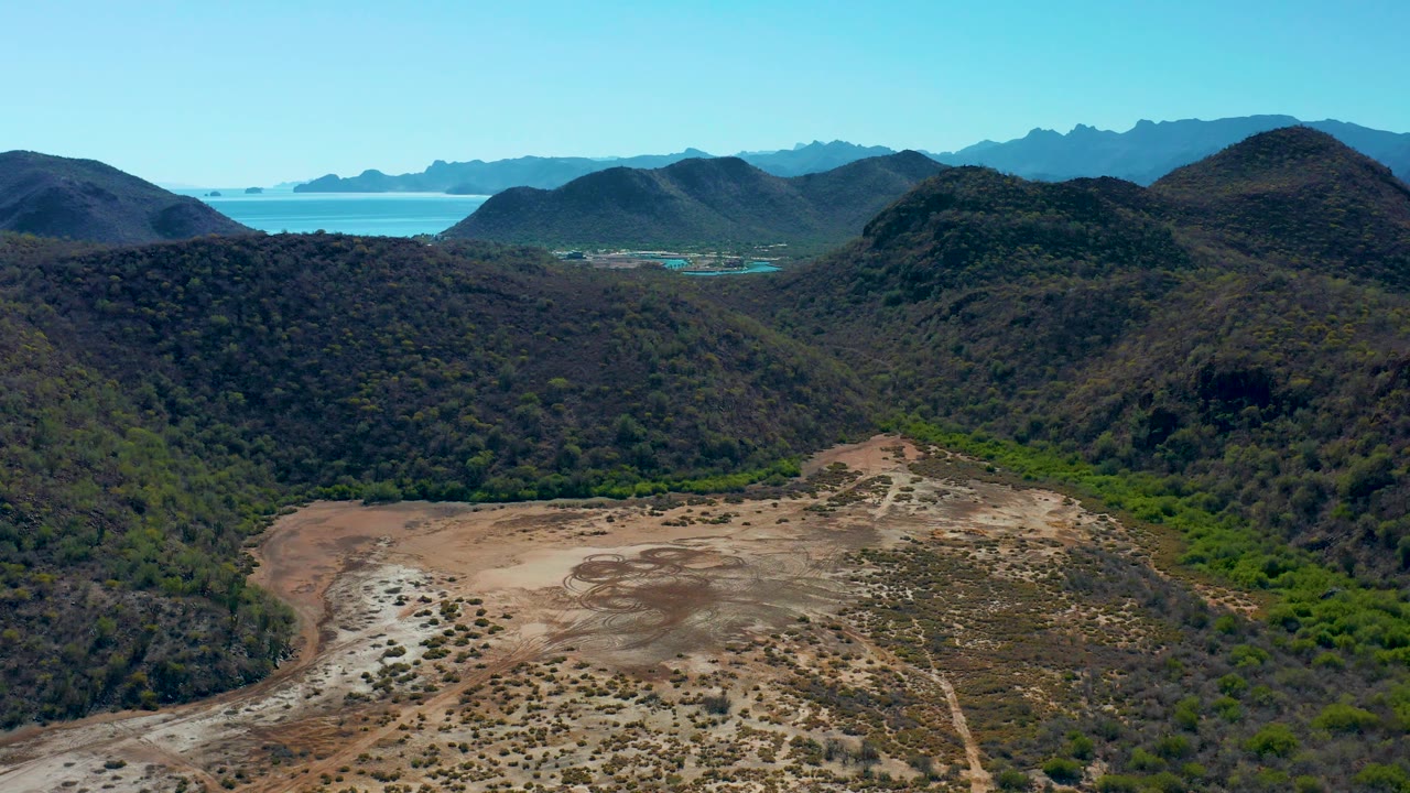 Loreto, Baja California Sur aerial landscape with dry foreground terrain, green vegetation in a wash, dark rounded hills, distant blue ocean, and hazy mountain layers, no visible people.