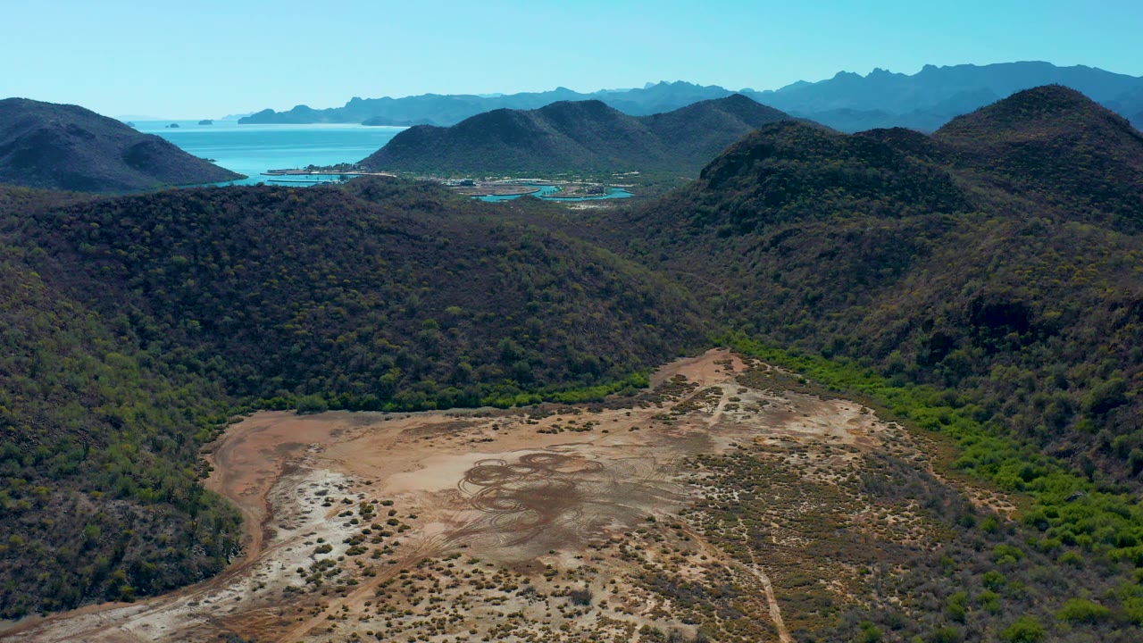 Early frame over Loreto, Baja California Sur, showing the tan riverbed area lower in frame, rounded green-brown hills in the middle distance, blue coastal water beyond, and no visible people.