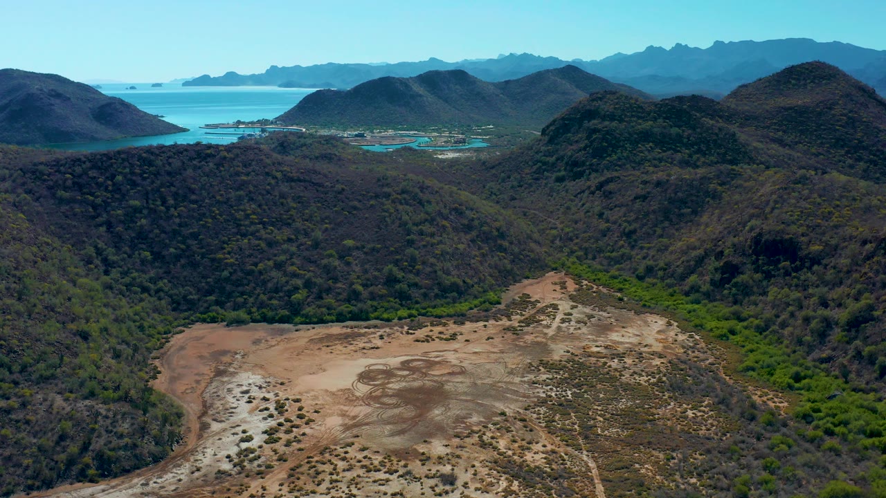 Aerial frame from the middle of the clip over Loreto, Baja California Sur, where the dry valley floor sits lower left, darker hills occupy the center, ocean water appears behind them, and no visible people are present.