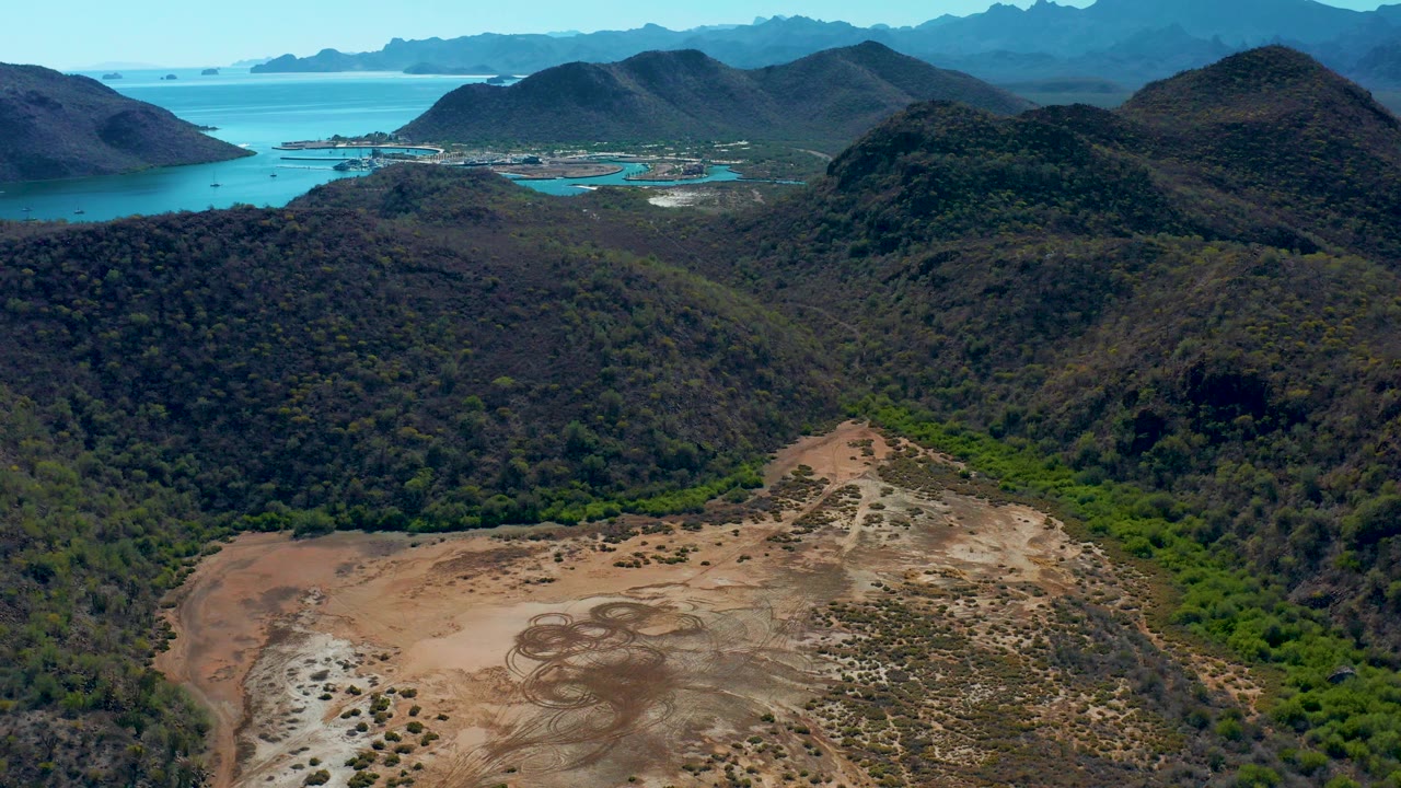 Opening aerial frame over Loreto, Baja California Sur with cleared brown terrain in front, green mountain slopes beyond, turquoise ocean at left, distant islands on the horizon, and no visible people.