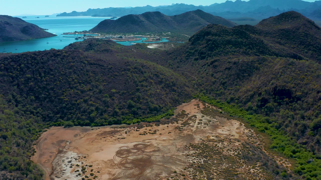 Wide aerial frame in Loreto, Baja California Sur showing a brown cleared area at the bottom edge, layered green and dark hills across the center, blue ocean and island shapes in the distance, and no visible people.