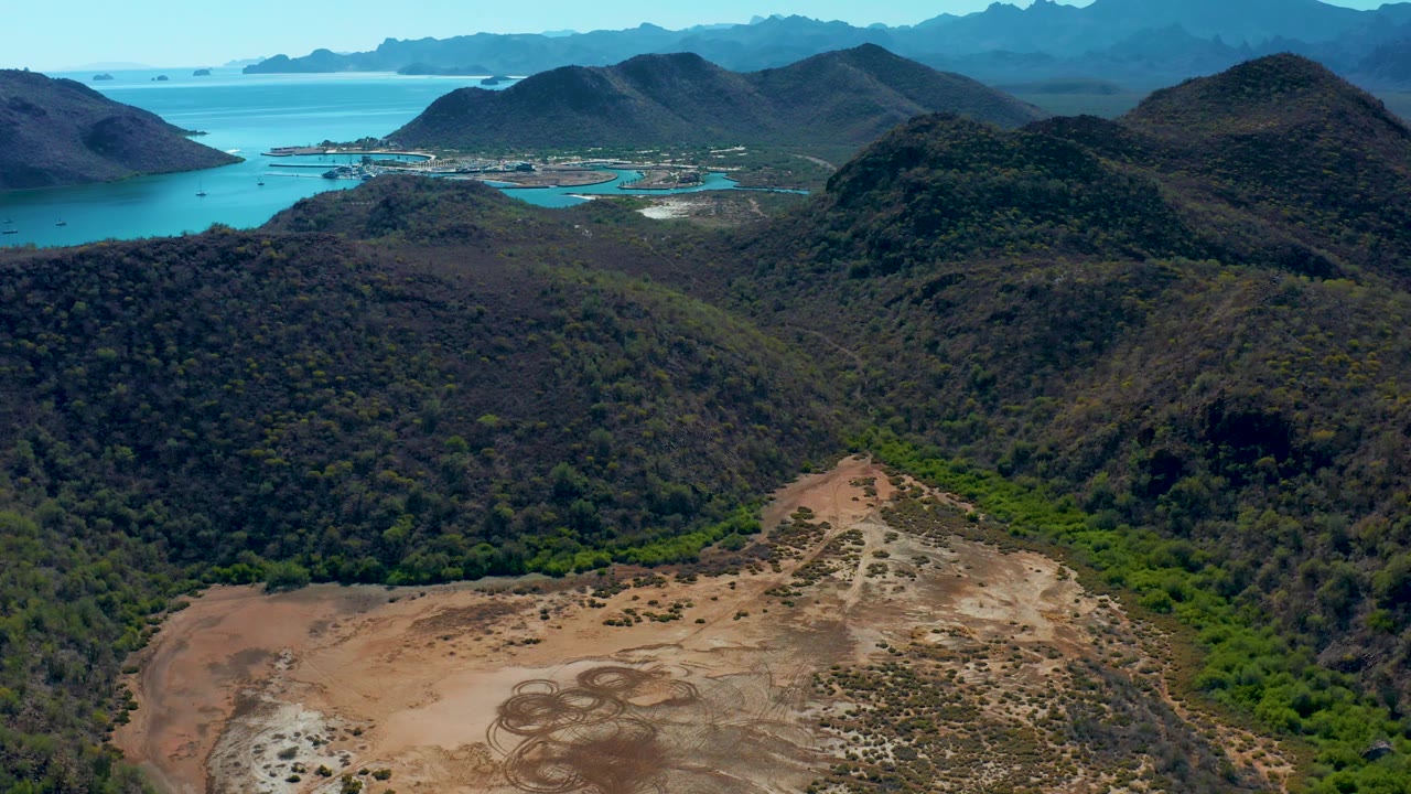 Aerial view of cleared valley floor below rugged hills and turquoise water.