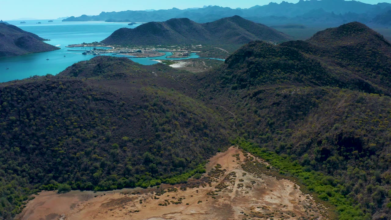 Drone view in Loreto, Baja California Sur, Mexico, with rugged hills in front, turquoise water beyond, a small shoreline settlement, and no visible people.