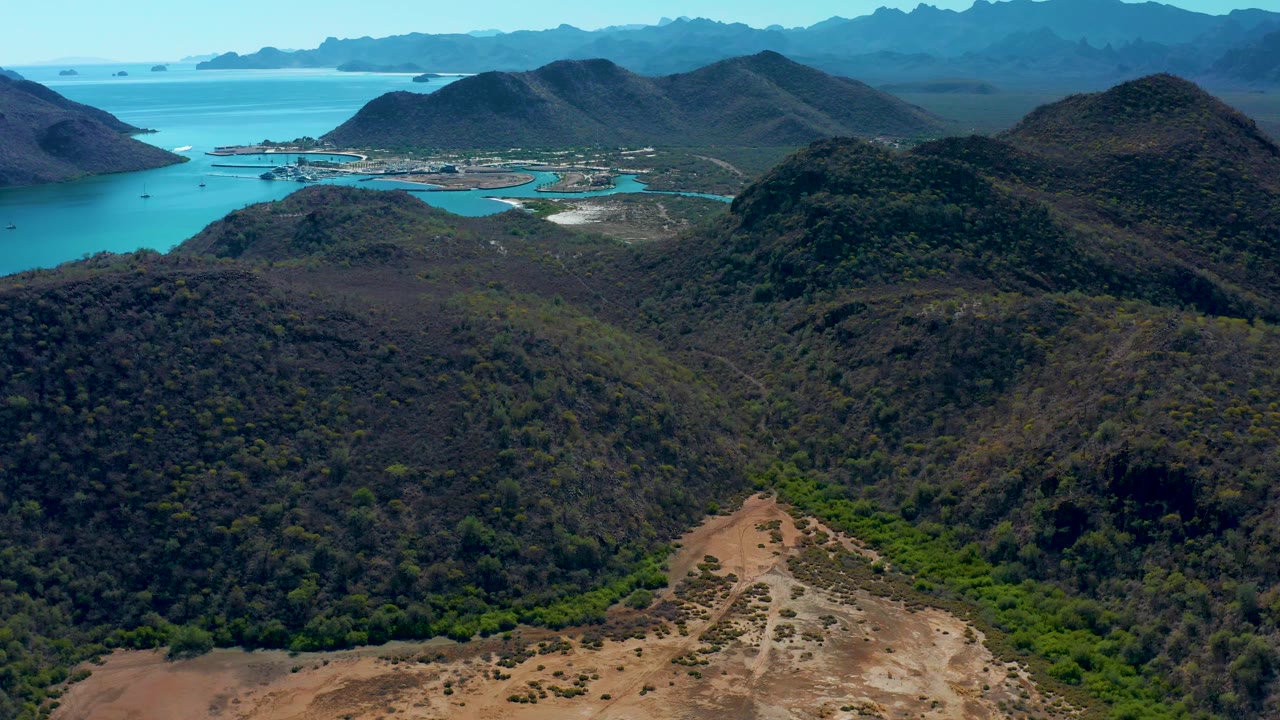 Aerial landscape with a dry foreground ridge, green valley, hills, and ocean beyond.