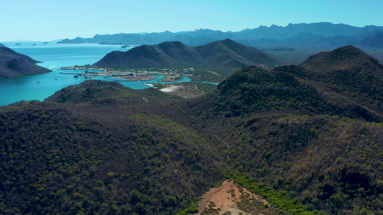 Aerial view over Loreto, Baja California Sur, with scrub-covered hills in front, turquoise bay water beyond them, distant mountain ridges under clear sky, and no visible people.