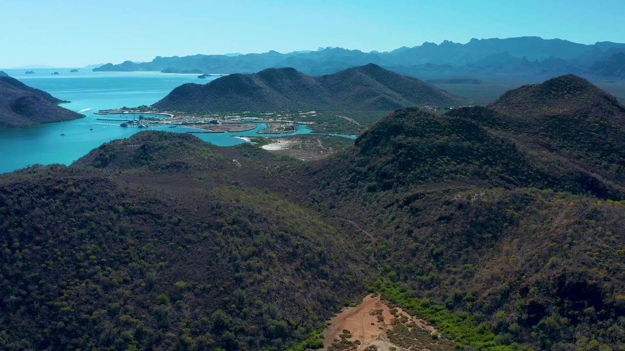 A high aerial frame in Loreto shows dark foreground hills, a pale exposed shore near the waterline, turquoise bay water, layered blue mountains, and no visible people.