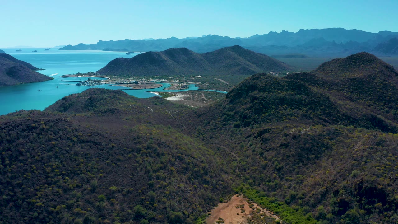The frame looks across a foreground ridge toward Loreto's bay, where bright water wraps around low landforms and blue mountain ranges recede into the background, with no visible people.