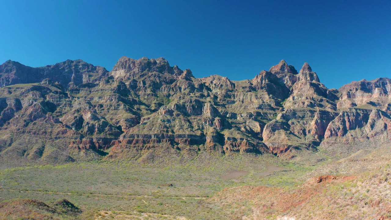 Opening view of stratified mountains above an arid Loreto, Baja California Sur valley with sparse vegetation, rocky foreground, clear blue sky, and no visible people.