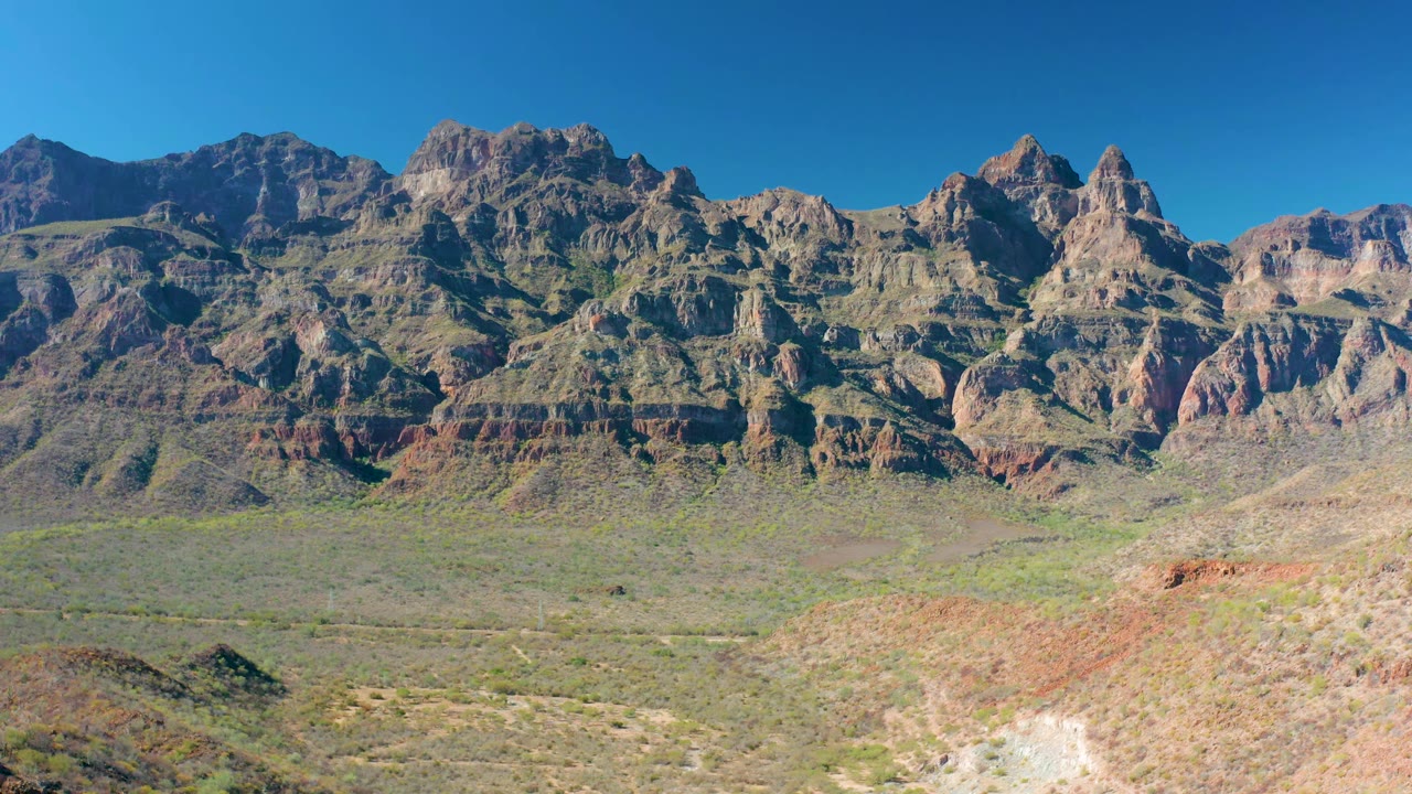Wide frame of Loreto mountains with jagged peaks, reddish rock layers, scrub-covered valley ground, bright daylight, and no visible people.