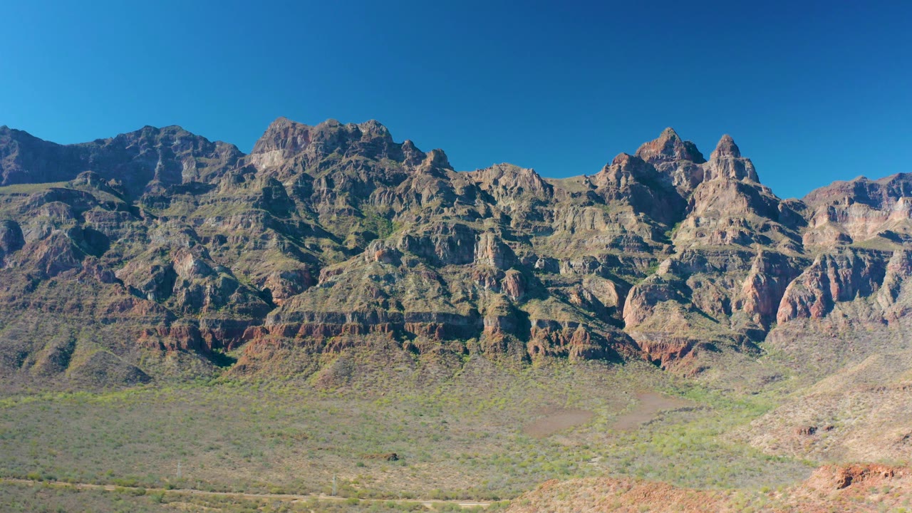 Layered stone ridges rise above pale arid ground in Loreto, with a cloudless blue sky and no visible people.