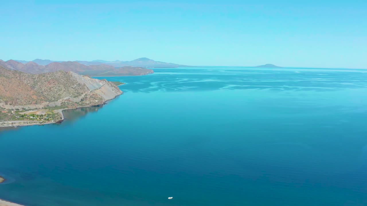 Wide aerial frame near Loreto, Baja California Sur, with rocky coast on the left edge, broad turquoise sea, faint water texture, distant mountains, and no visible people.