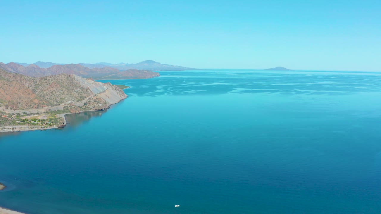 Aerial view over Loreto coastal water, with the left shoreline receding into dry hills, a small white boat low in frame, distant landforms, clear sky, and no visible people.