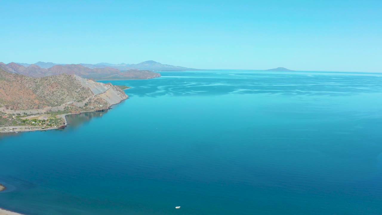 Aerial frame near Loreto, Baja California Sur, showing the rocky left shoreline, calm turquoise water, distant mountains, pale blue sky, and no visible people.