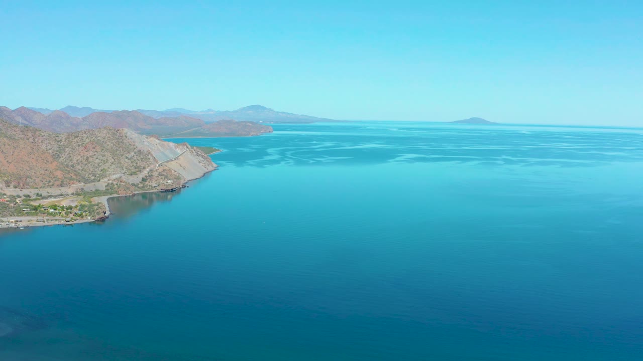 Aerial view over Loreto with turquoise water filling the lower frame, dry hills and a pale rocky point on the left, faint wave patterns offshore, and no visible people.